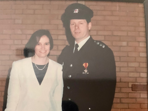 Tom & Una in their late 20s/early 30s. Una is wearing a cream blazer and striped black and grey blouse. Tom is wearing his official fireman's uniform.